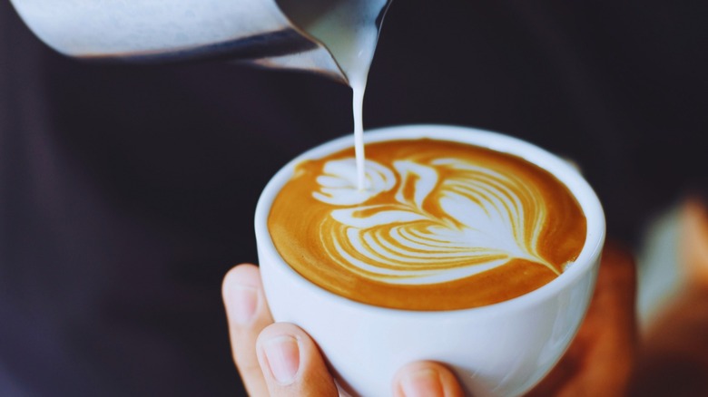 Person pouring steamed milk into latte in white cup